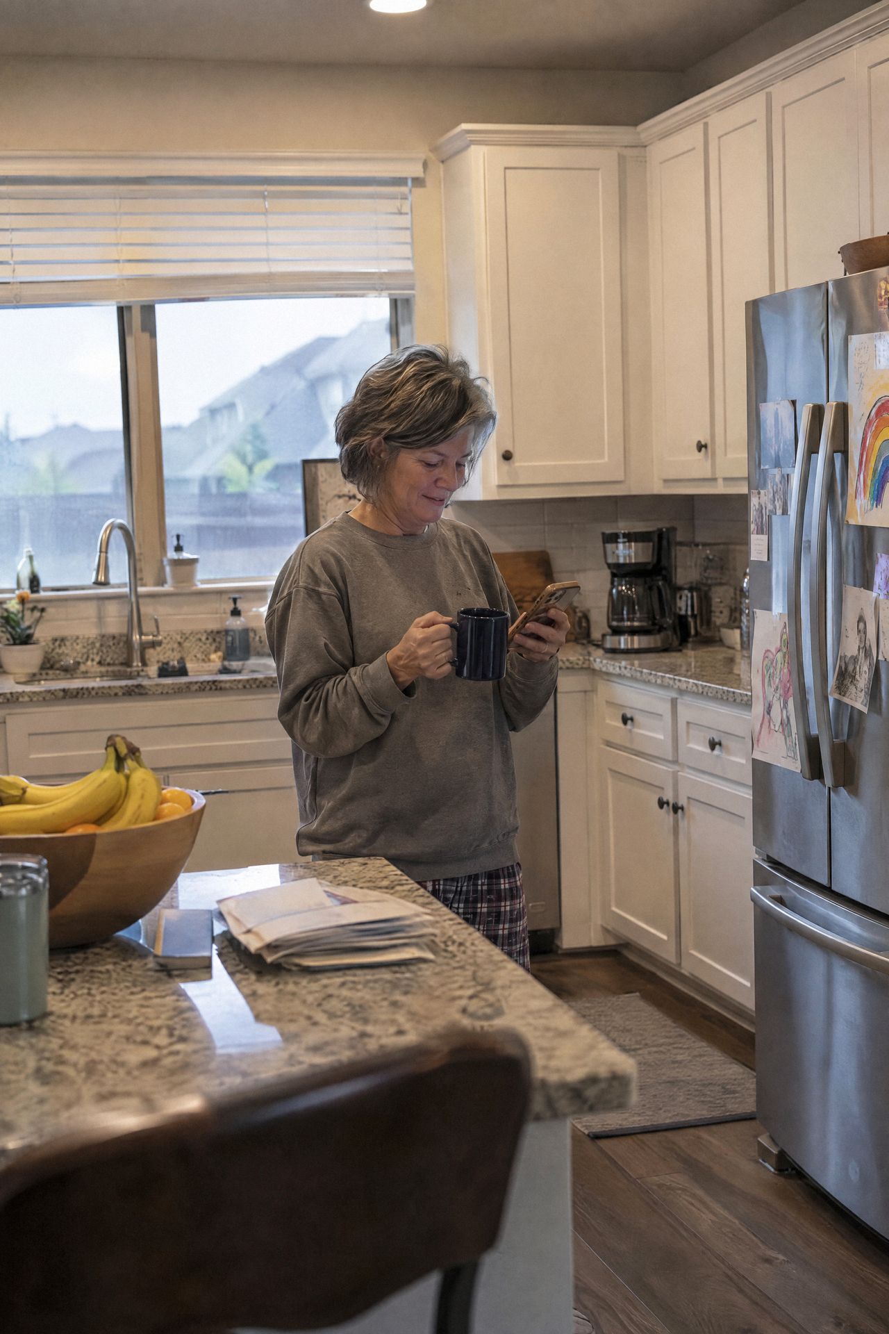 A woman in her kitchen reads her phone with a coffee mug in hand